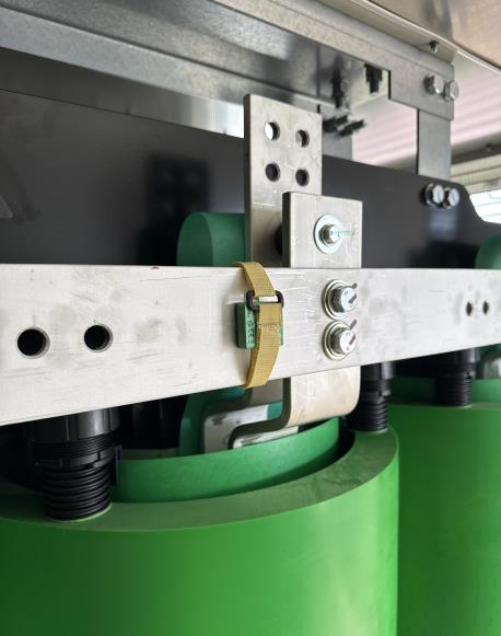 Close-up of industrial machinery showing green rollers, a metal frame with bolts, and a yellow strap fastened around one part. The setting appears to be indoors with some light coming from the right.
