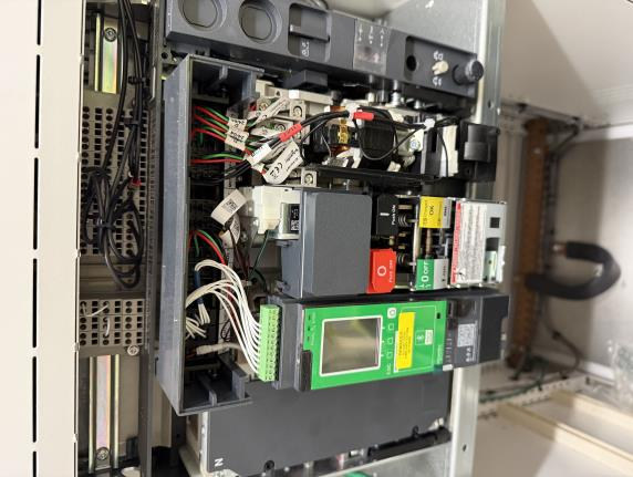 Close-up of an open electrical control panel showing various components, wires, labels, and circuit breakers, including a green Schneider Electric device with a display screen and buttons. Panel is vertically oriented.