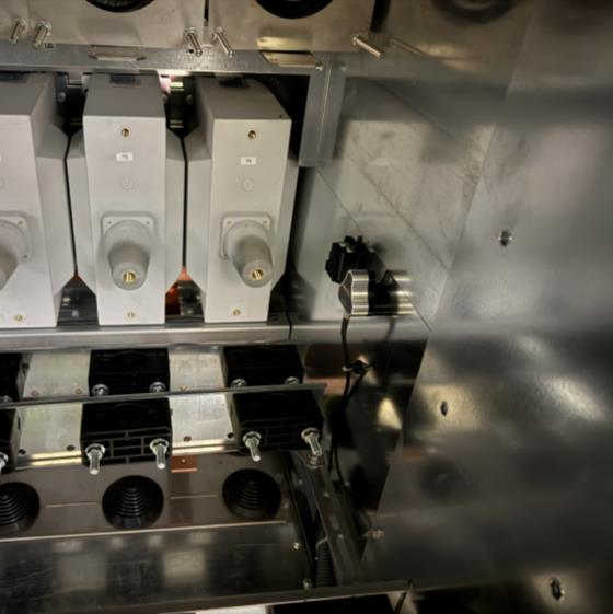 Close-up view of electrical components inside a metallic enclosure, featuring three prominent connectors or fuses, various black cables, and shiny metallic surfaces with reflections.