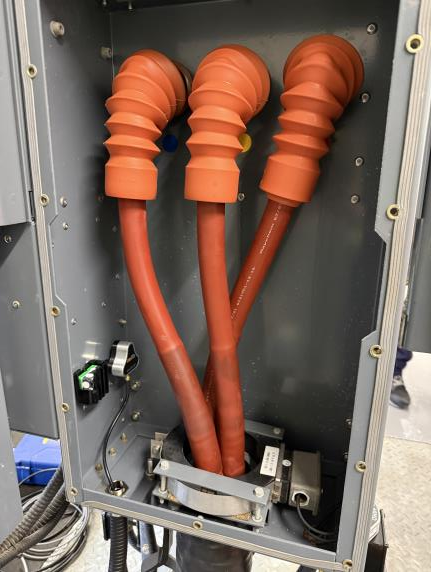 Three thick orange insulated cables run into a metal electrical enclosure, each fitted with a flexible rubber boot where they enter. The inside contains some wiring, components, and connectors.