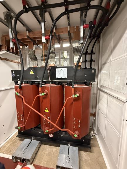 Large electrical transformer with three cylindrical coils, connected by thick black and copper wires, mounted on a black base inside an industrial room with white paneled walls and metallic components visible.
