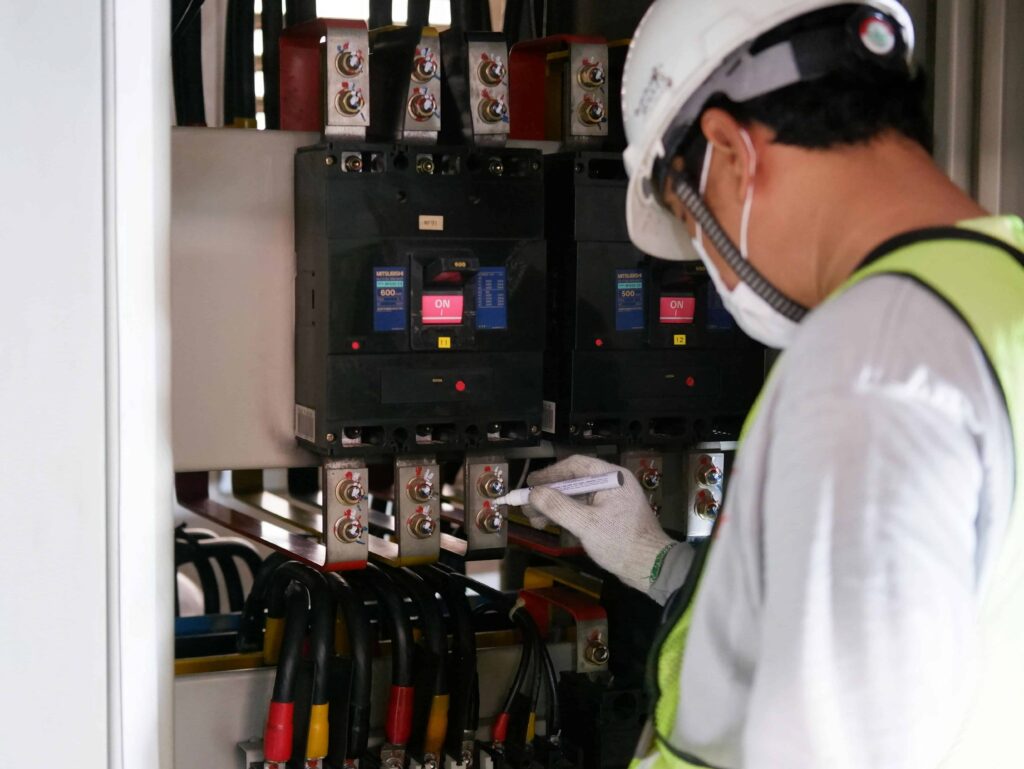 A worker wearing a helmet, mask, gloves, and safety vest writes notes while inspecting electrical panels with wires and switches in an industrial setting.