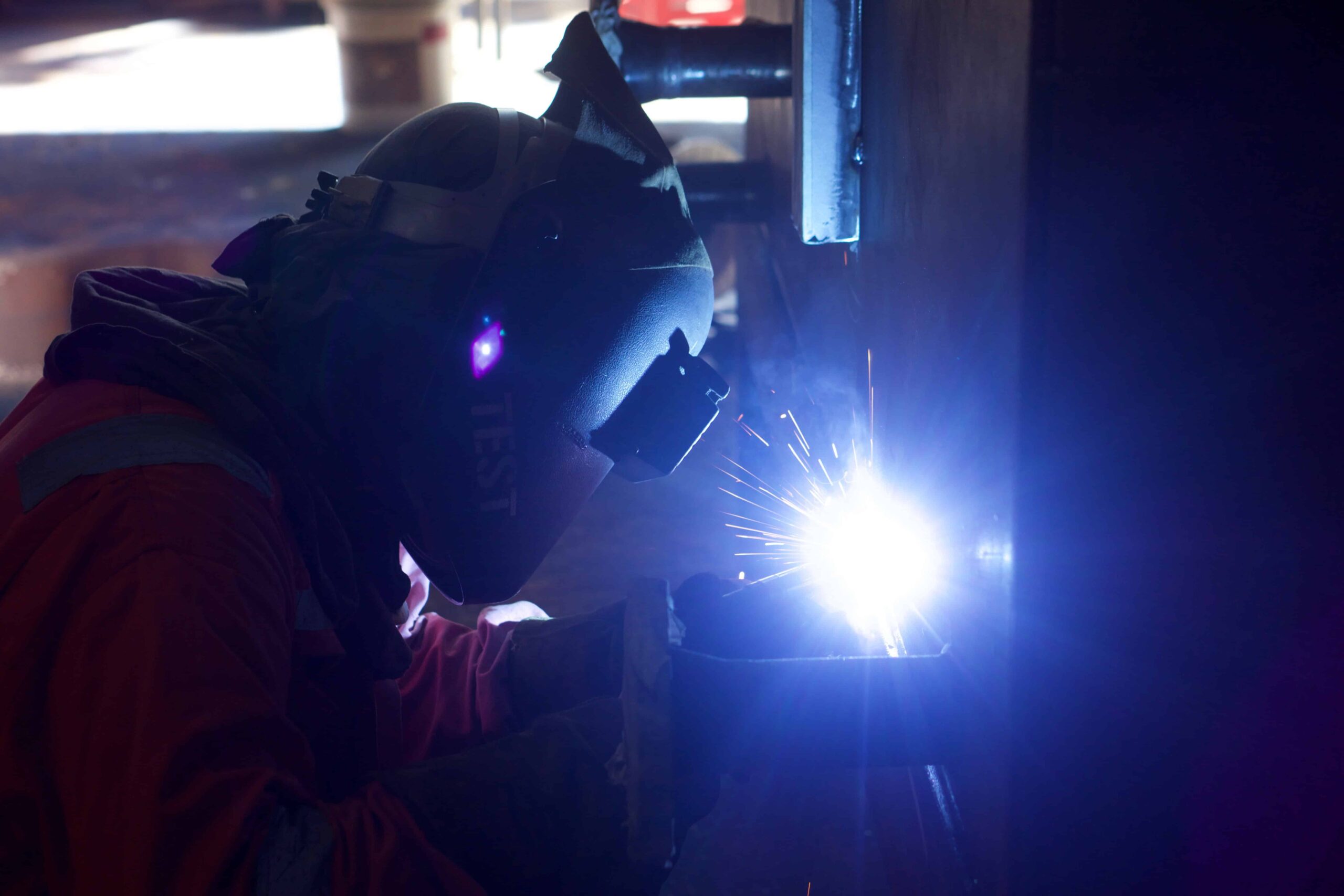 A person wearing protective gear and a welding helmet is welding metal, with bright sparks and light visible at the point of contact. The worker appears to be indoors in a dimly lit area.